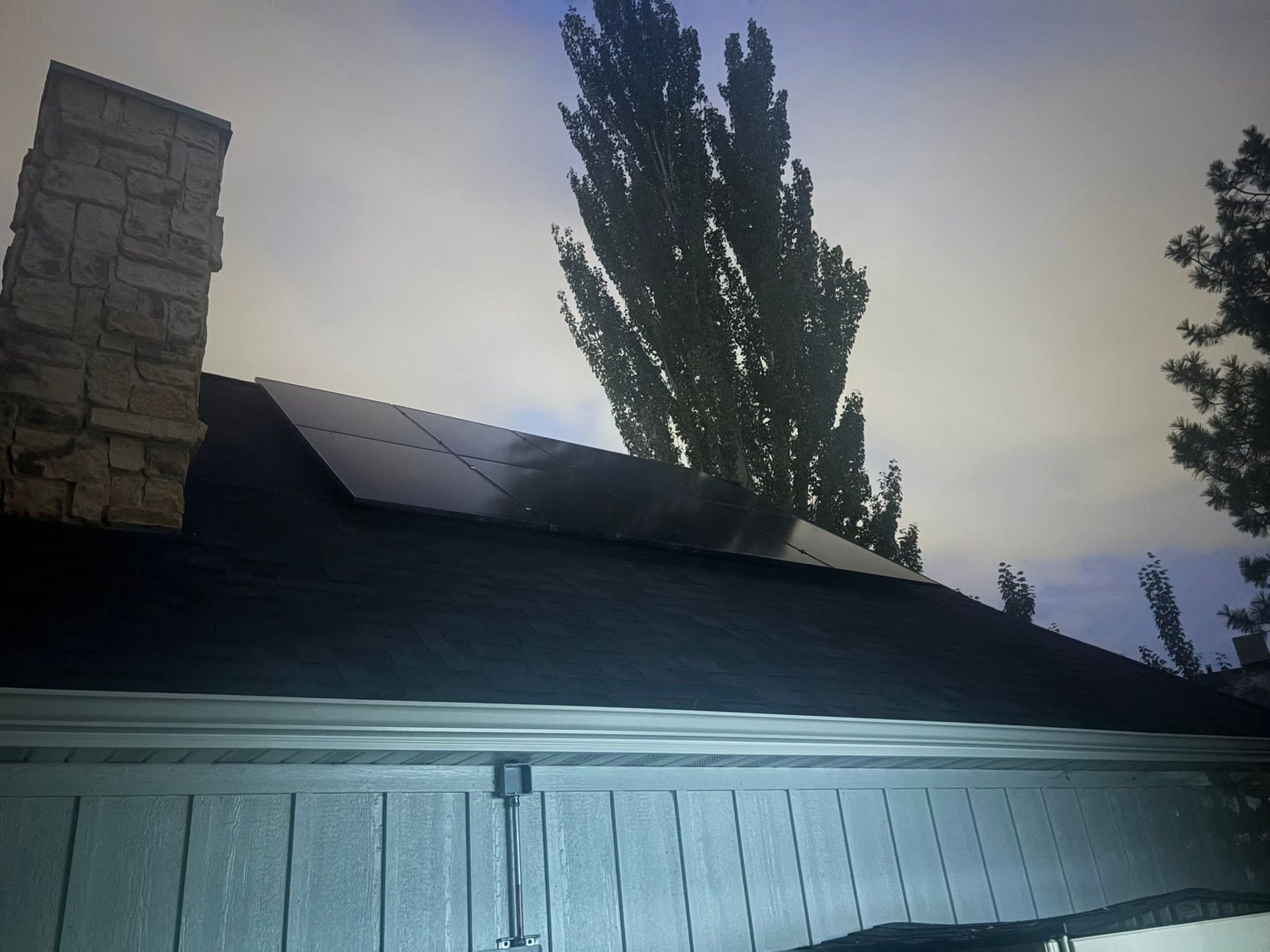 Rooftop with solar panels beside a stone chimney, tall tree, and bright sky at dusk