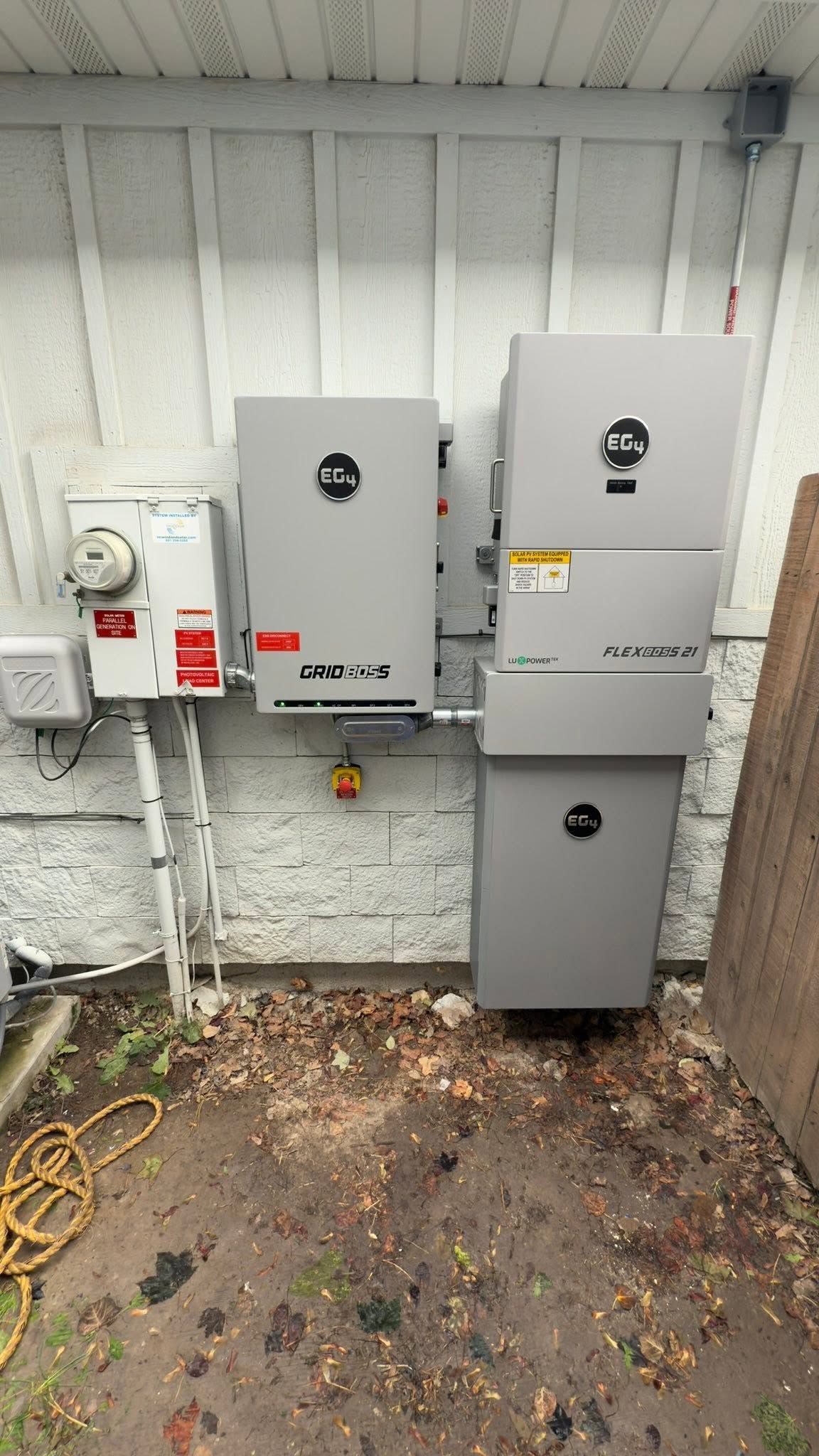 Two gray electrical service panels mounted on an exterior wall beside conduit and wet ground.