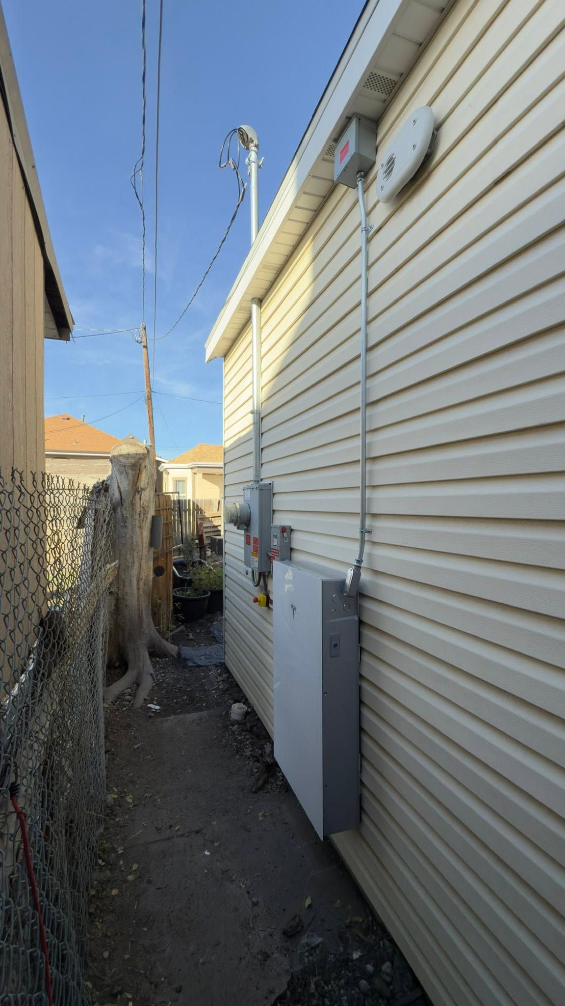 Narrow side yard between houses with dirt ground, chain-link fence, and house utilities on the right wall