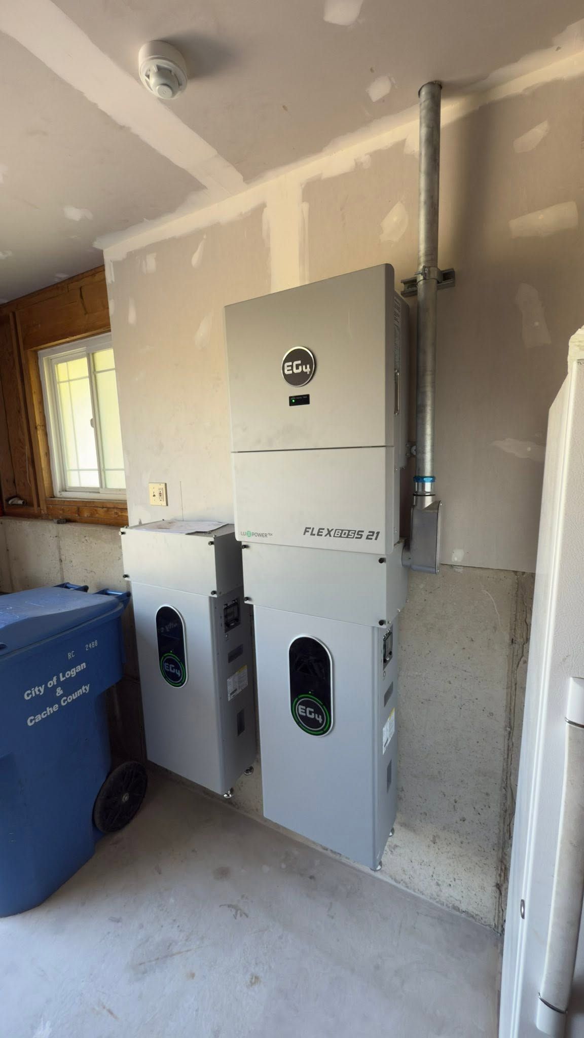 Utility meters mounted on a garage wall beside a blue recycling bin, with unfinished drywall around them