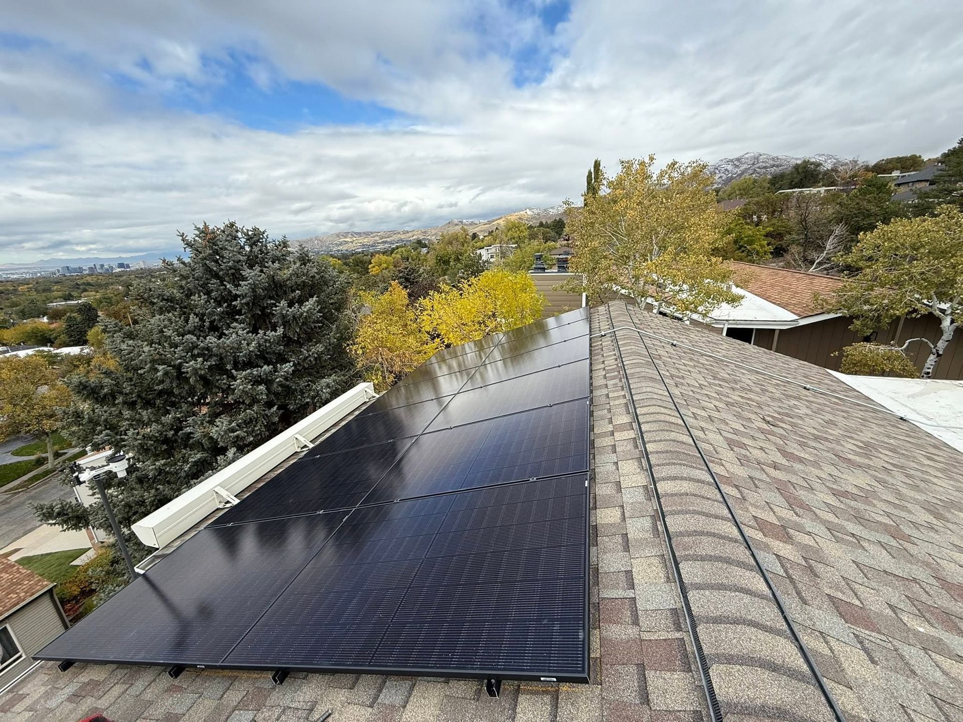 Solar panels on a rooftop beside shingled roof tiles and autumn trees under a cloudy sky