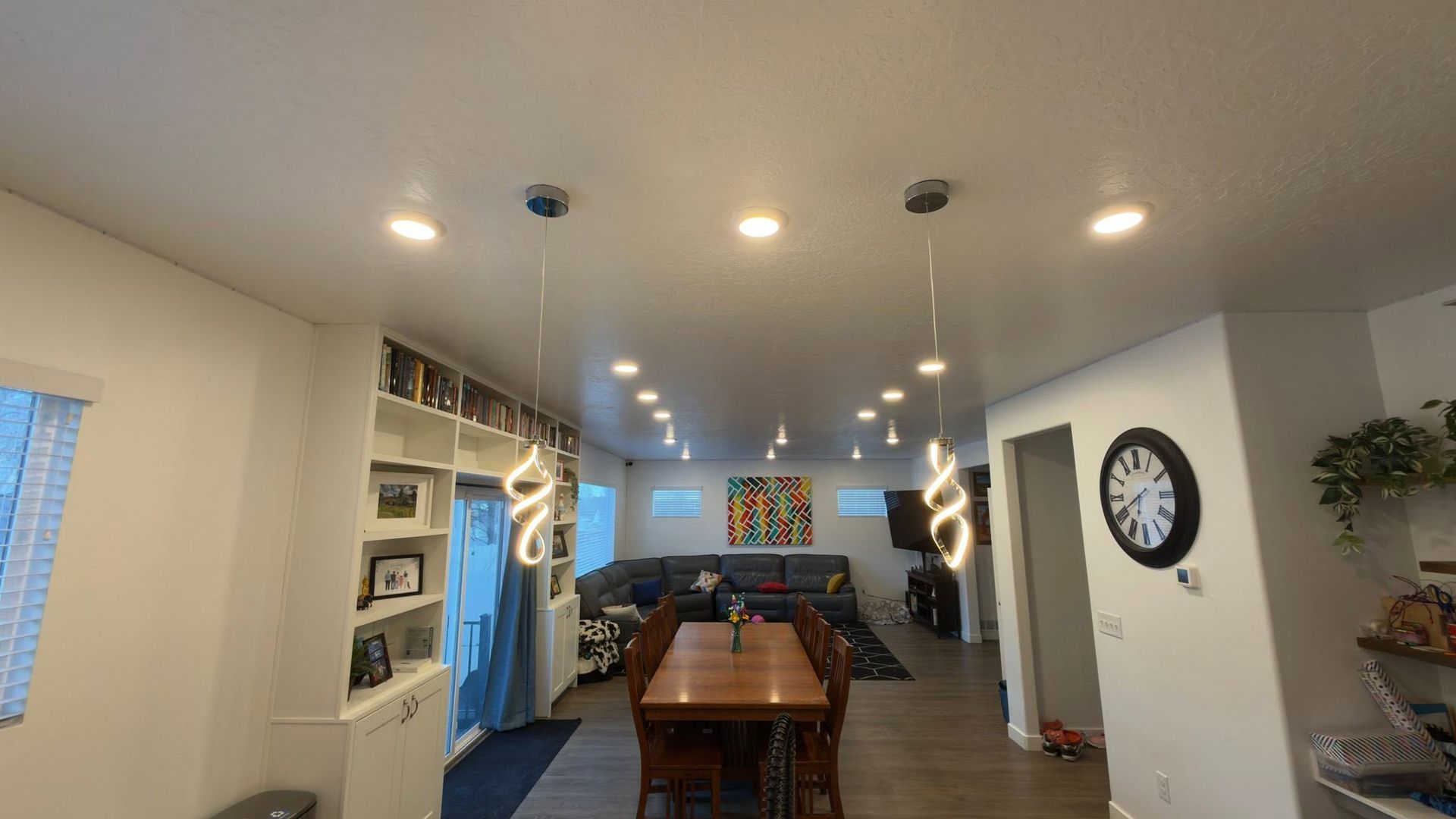 Bright open kitchen and dining area with recessed lights, wood table, wall clock, and shelves.