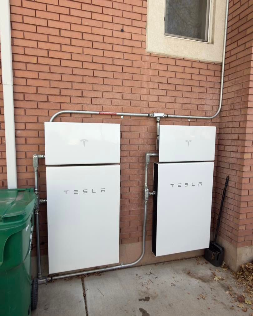 Two white Tesla Powerwall batteries mounted on a brick exterior wall with conduit piping