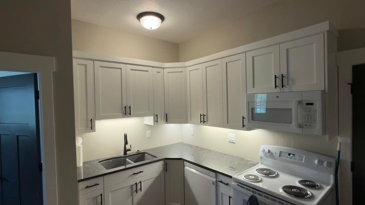 Compact white kitchen with white cabinets, stove, microwave, sink, and under-cabinet lighting