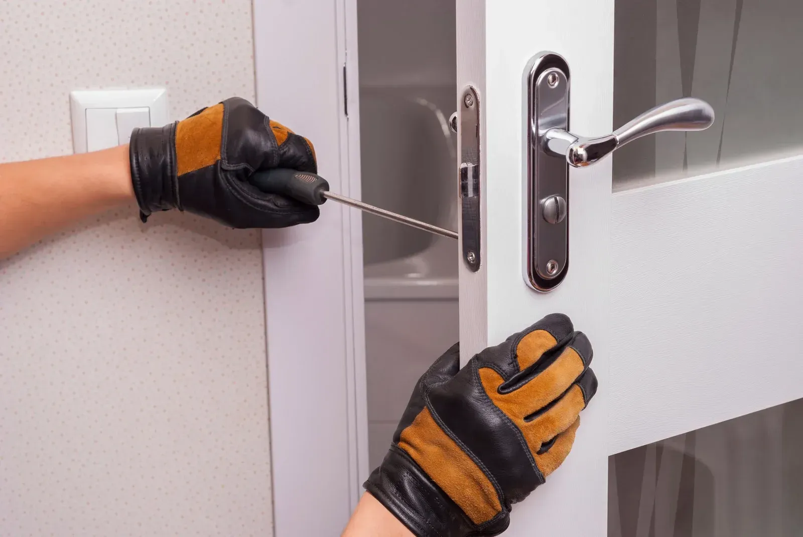 Gloved hands using a screwdriver to adjust a silver door lock on a white door