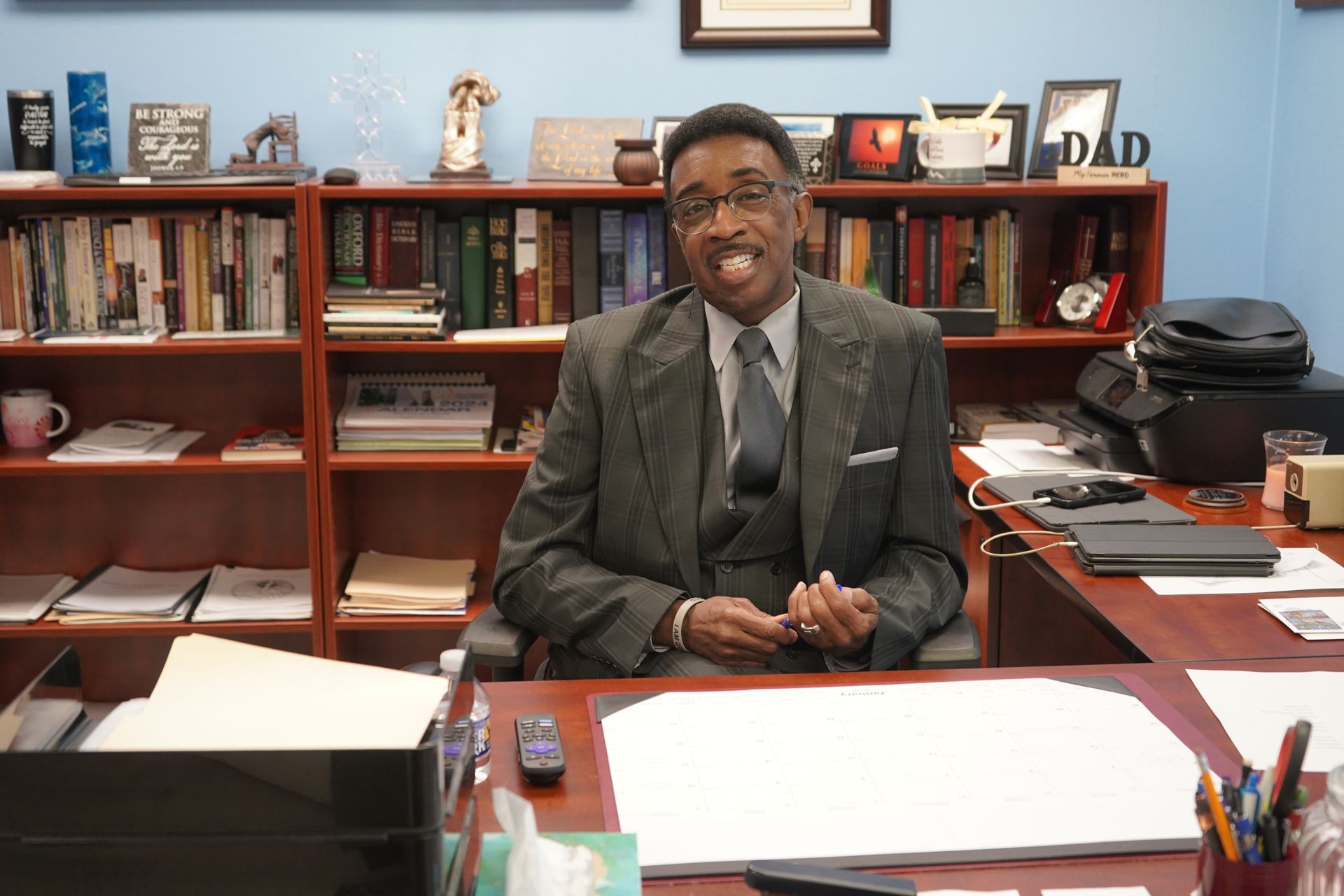 A man in a suit and tie sits at a desk in an office with bookshelves in the background, smiling at the camera.