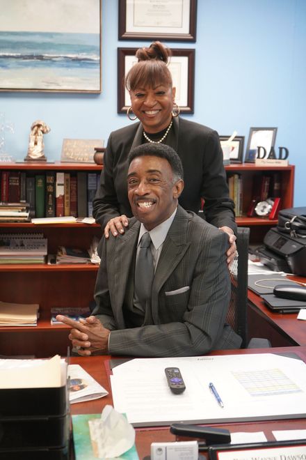 A professional couple smiling in an office, with a man seated at a desk and a person standing behind him with a hand on his.