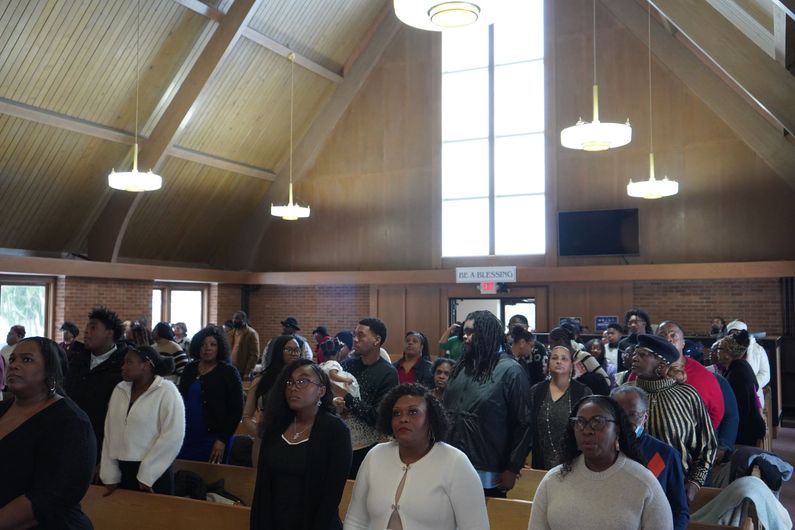 A diverse group of people stand in the pews of a church with wooden vaulted ceilings and hanging lights.