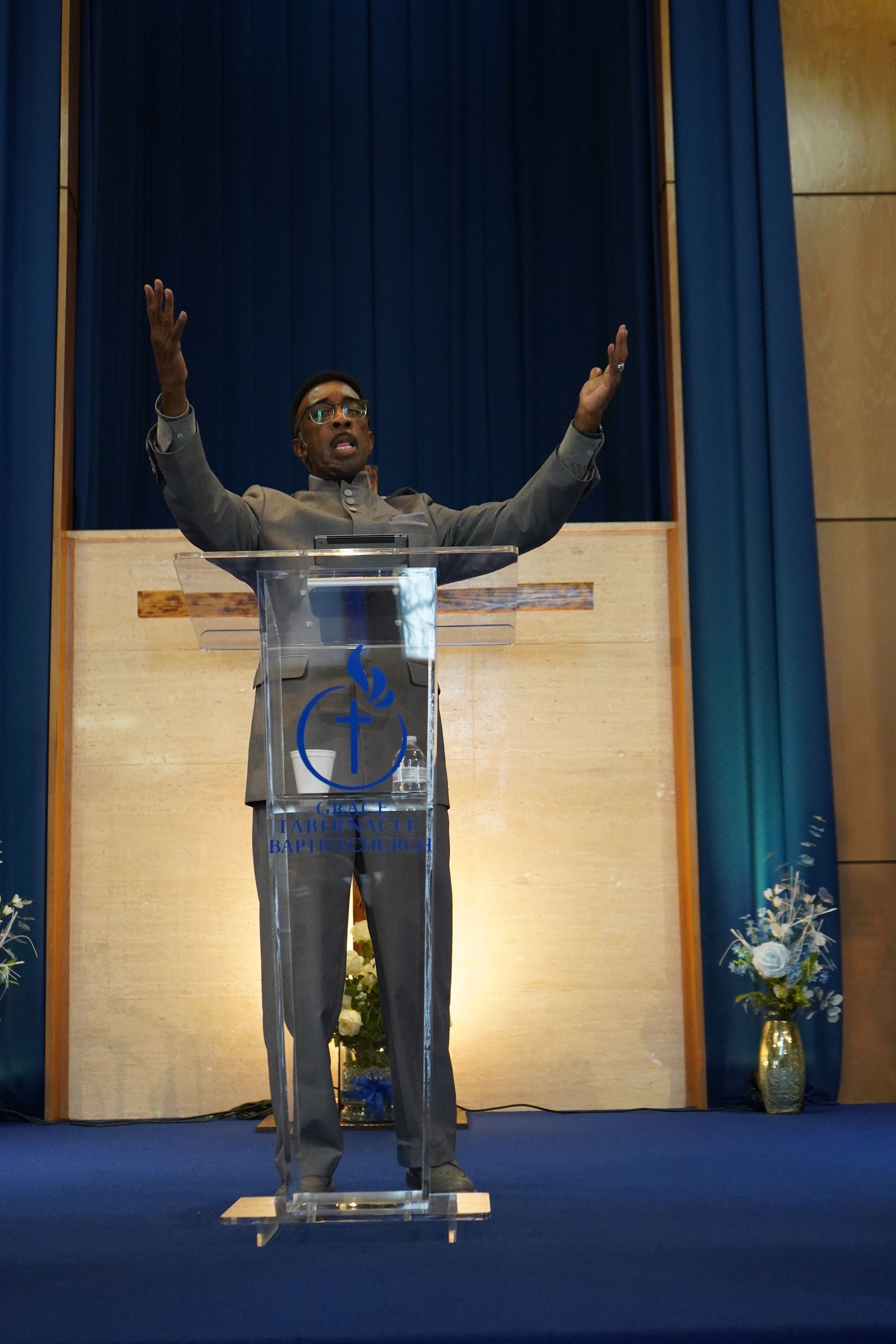 A person in a suit stands behind a clear podium, arms raised, speaking to an audience on a stage with blue curtains.