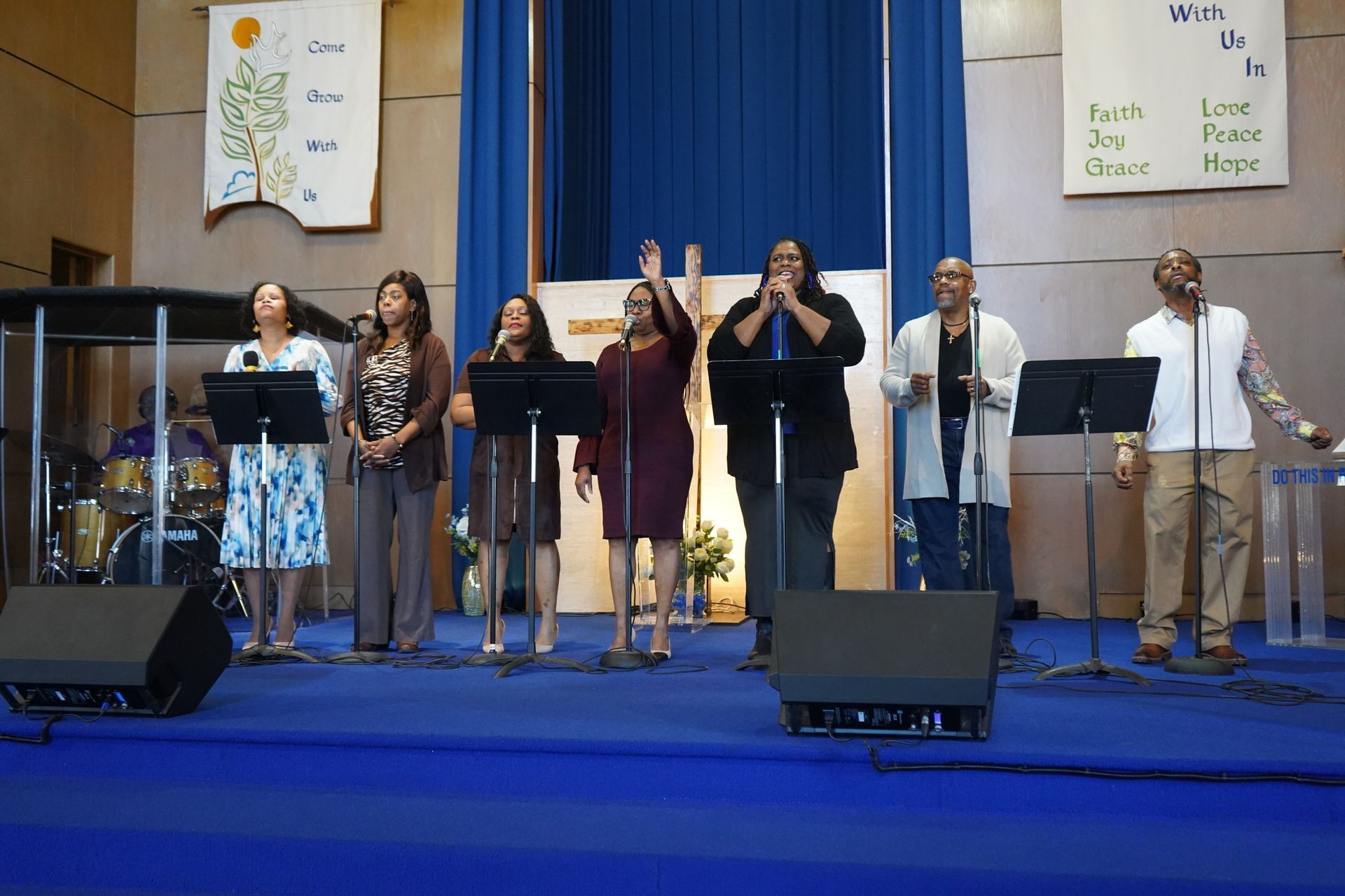 A musical group stands on a blue stage behind microphones and music stands, singing in a bright church hall.
