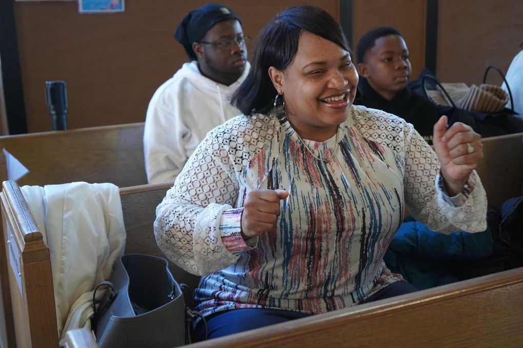 A smiling person sits in a wooden pew, gesturing with raised hands while others sit nearby in a church-like setting.