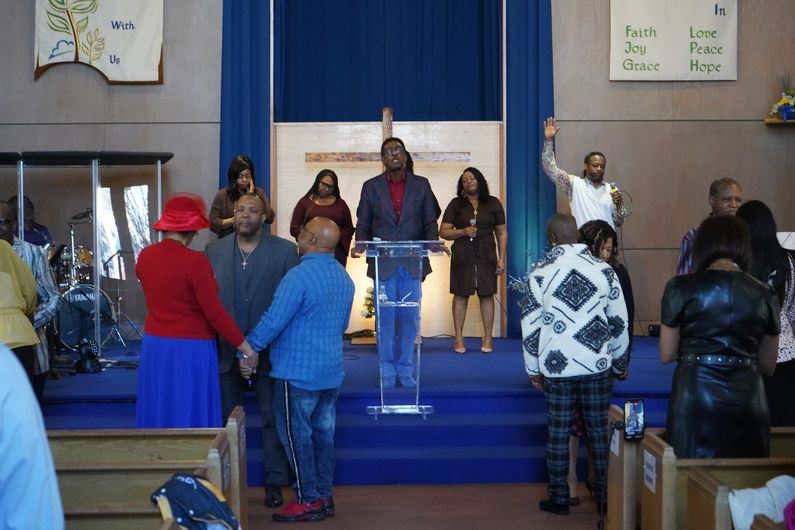 A congregation gathers in a church with a podium, blue carpet, and a cross on the wall as people stand and pray.