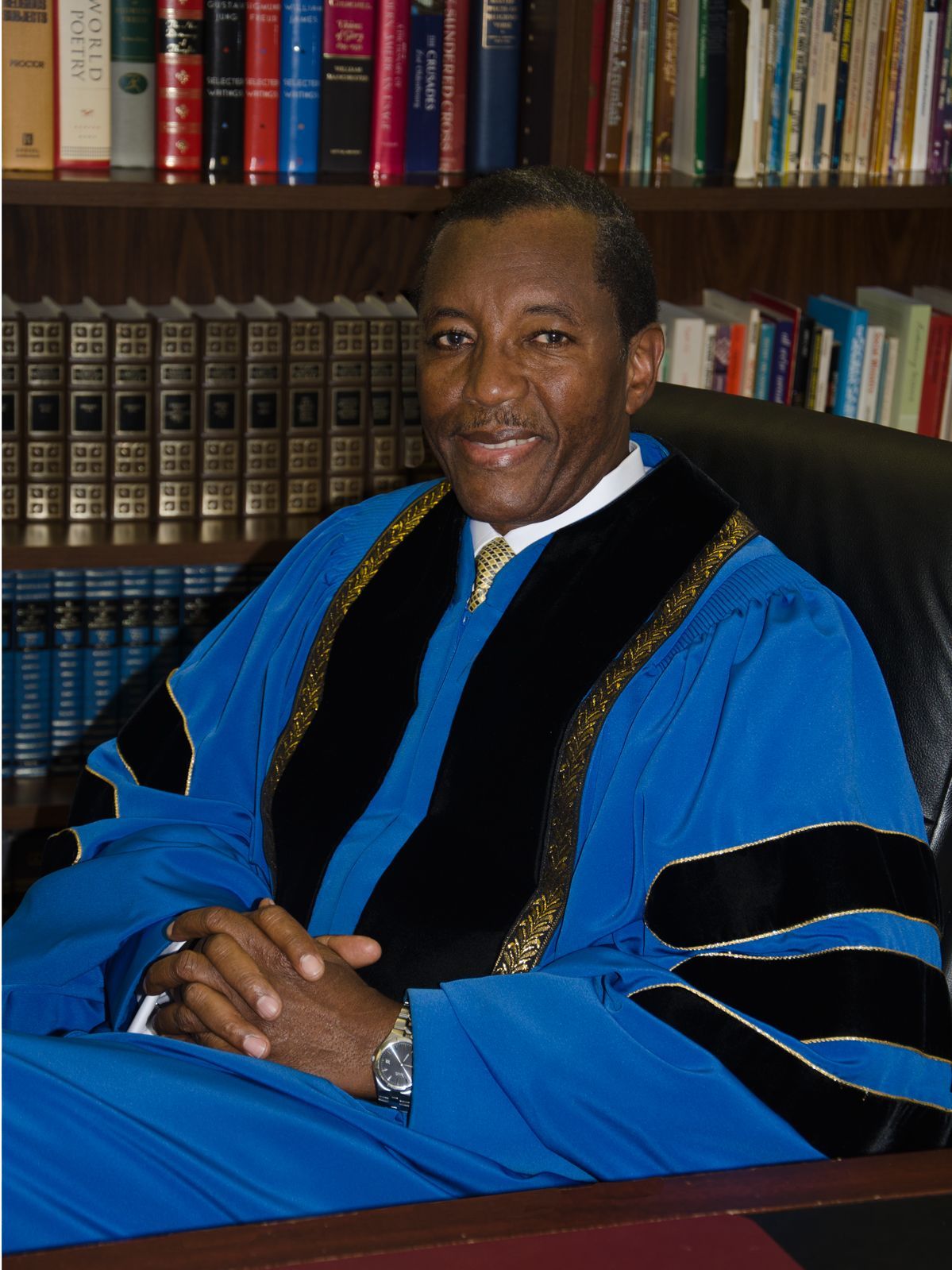 A person in a blue academic robe with black velvet panels and gold trim sits in a leather chair before a bookshelf.