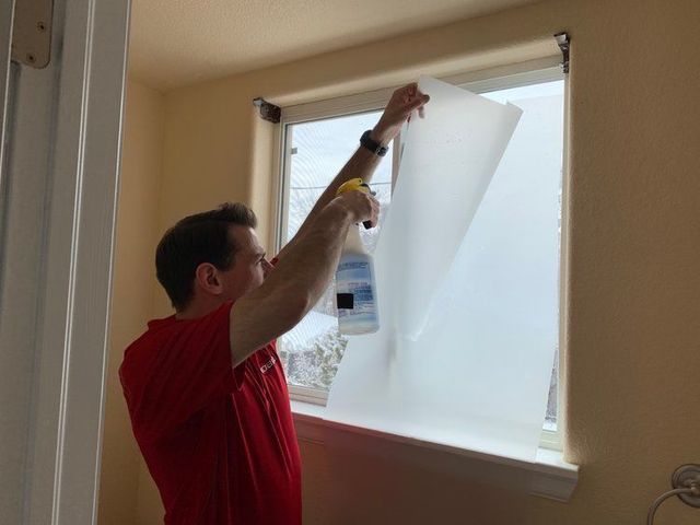 A man in a red shirt is covering a window with a white sheet of paper.