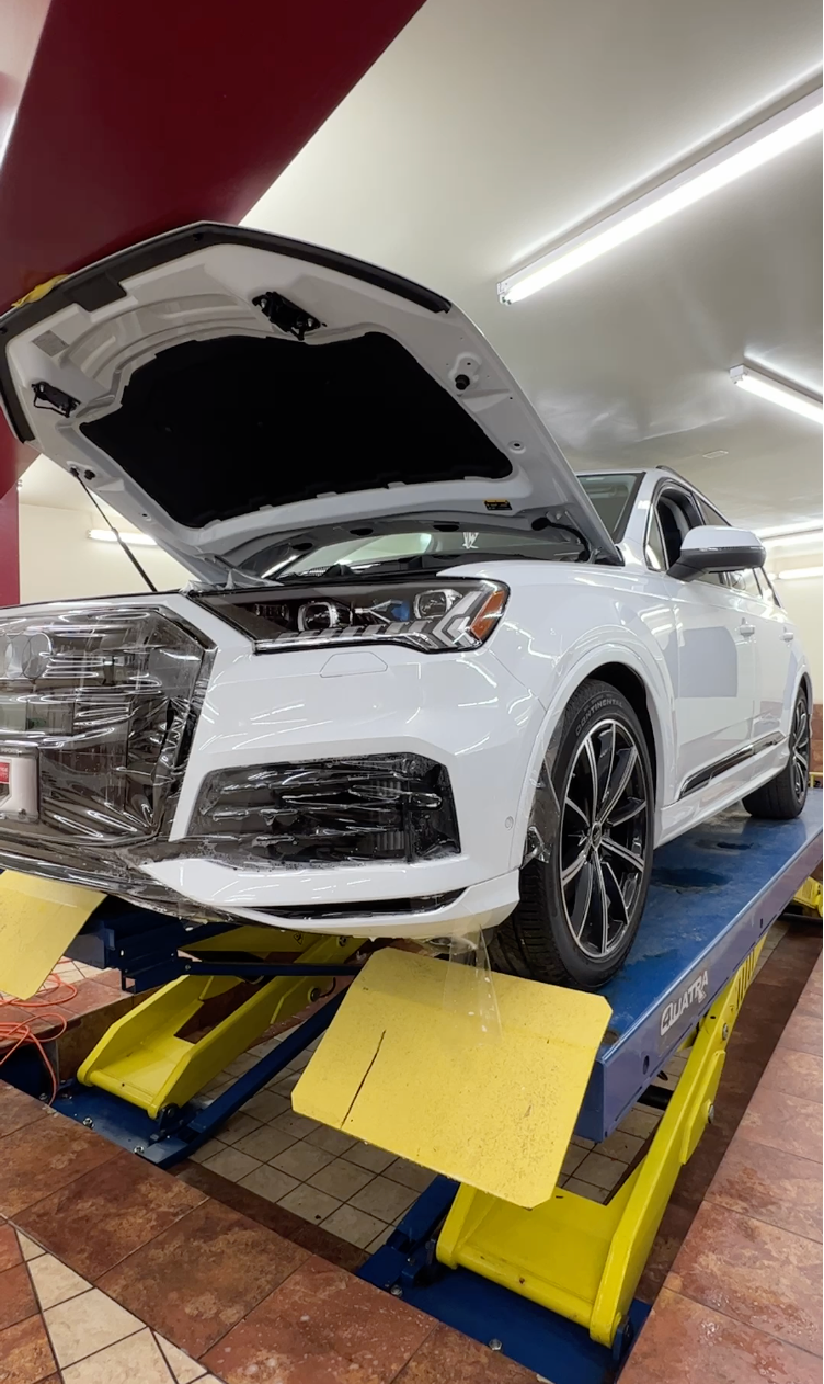 A white car is sitting on a lift with its hood open while getting a tint installation service.
