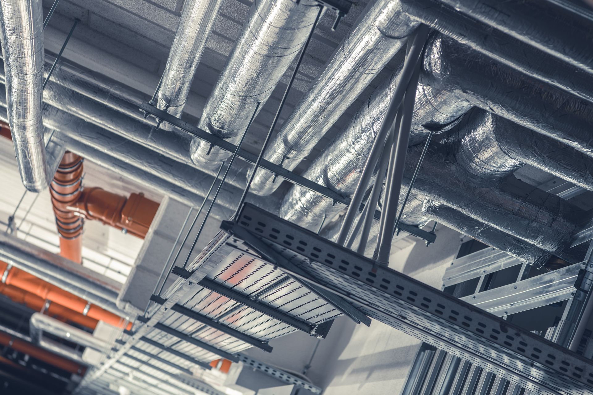 Industrial ceiling with exposed silver ducts, black cable trays, and orange pipes.