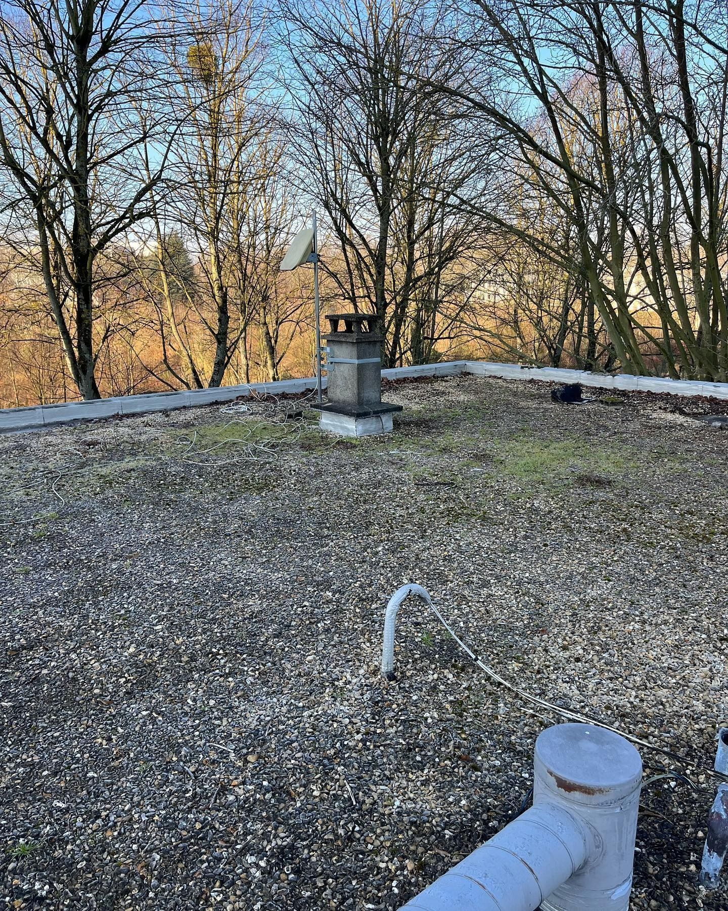 Toit plat en gravier avec cheminée et arbres dénudés en arrière-plan, sous un ciel bleu.