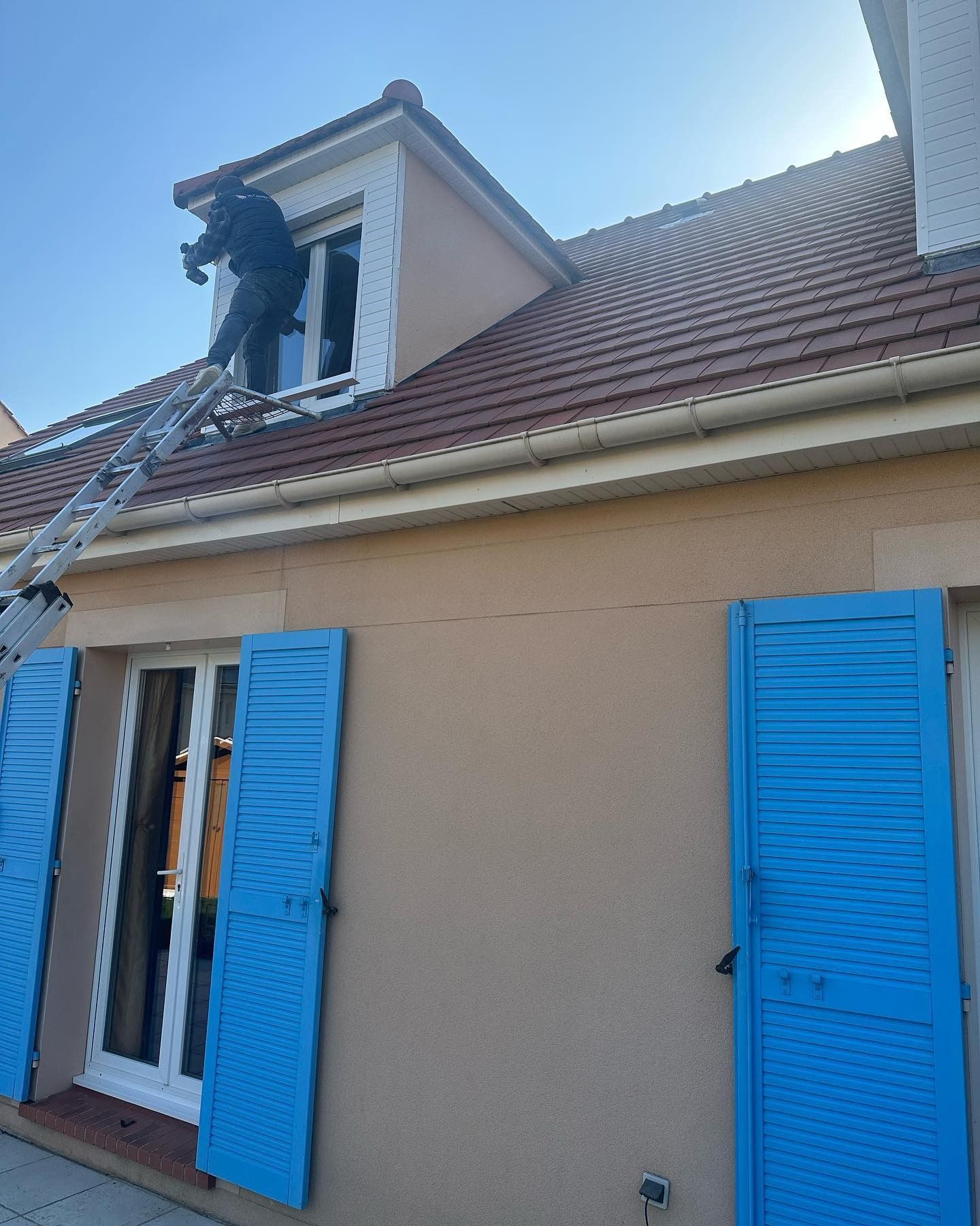 Bâtiment blanc dans le 78 avec une bâche bleue sur une terrasse en bois, une personne sur le toit.