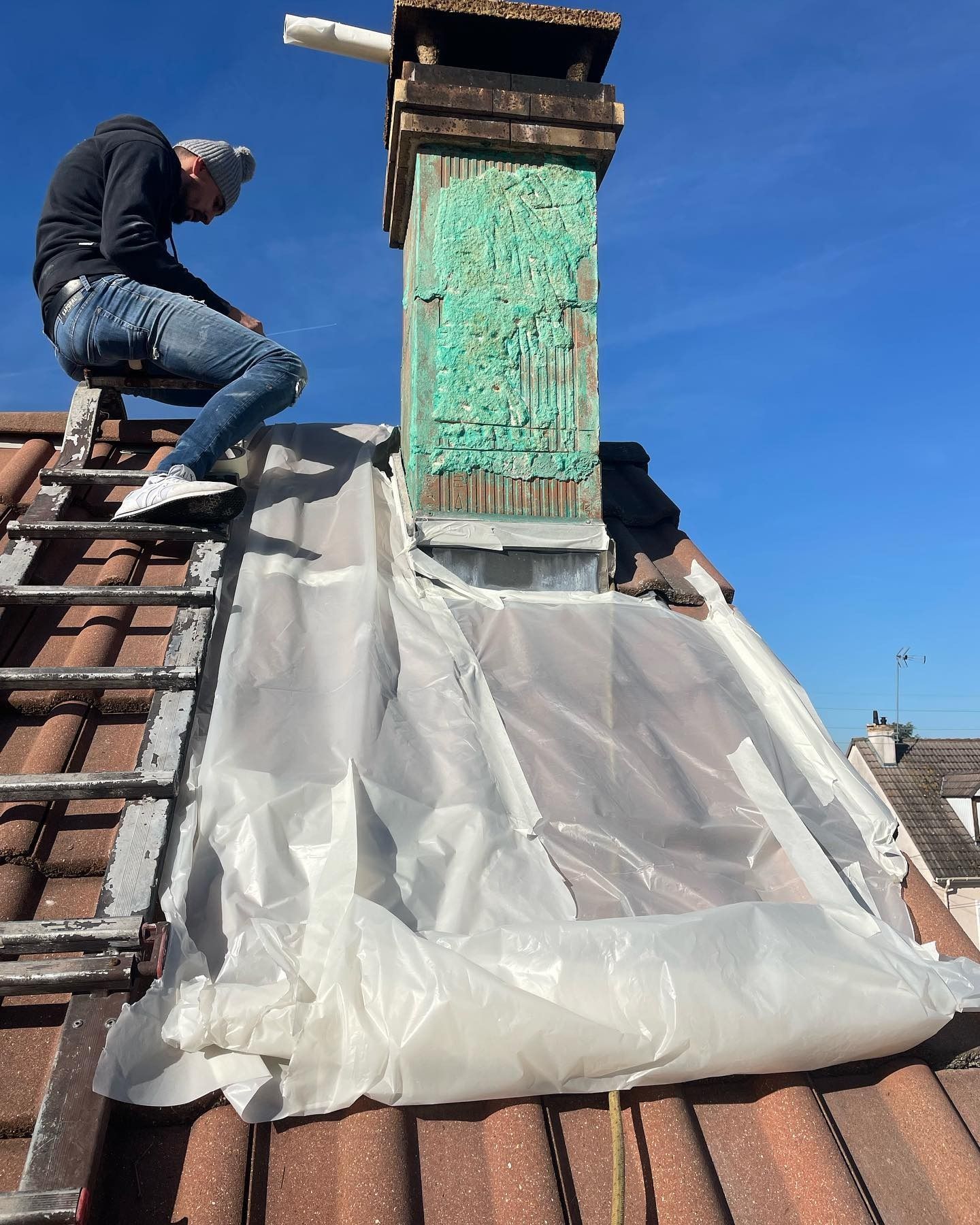 Une personne sur une échelle travaille sur une cheminée. Une bâche en plastique blanc recouvre le toit ; ciel bleu.
