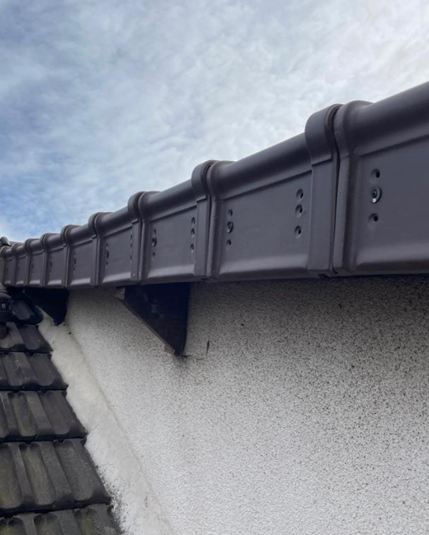 Gouttière brune fixée à un mur en stuc surmonté d'un toit de tuiles, ciel nuageux en arrière-plan.