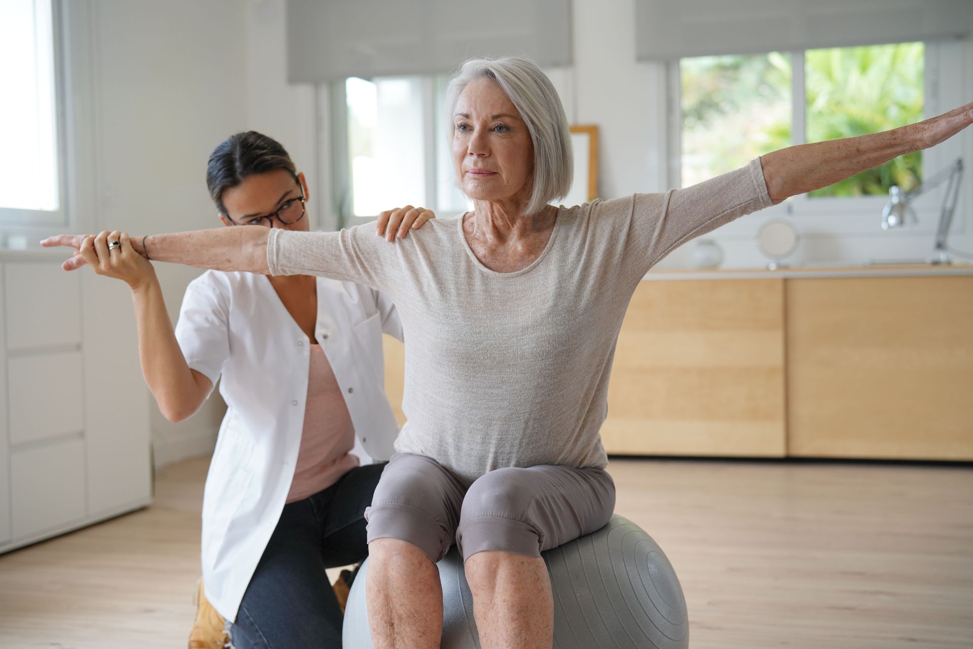 Eine ältere Frau sitzt auf einem Gymnastikball, während ihr eine Krankenschwester hilft.