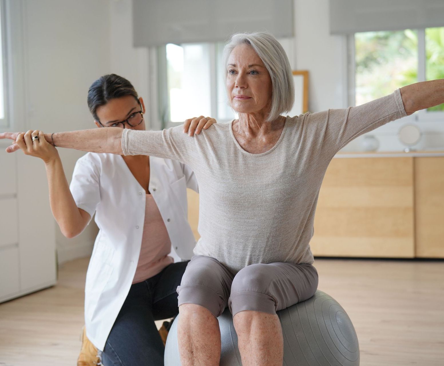 Eine ältere Frau sitzt auf einem Gymnastikball, während ihr eine Krankenschwester hilft.