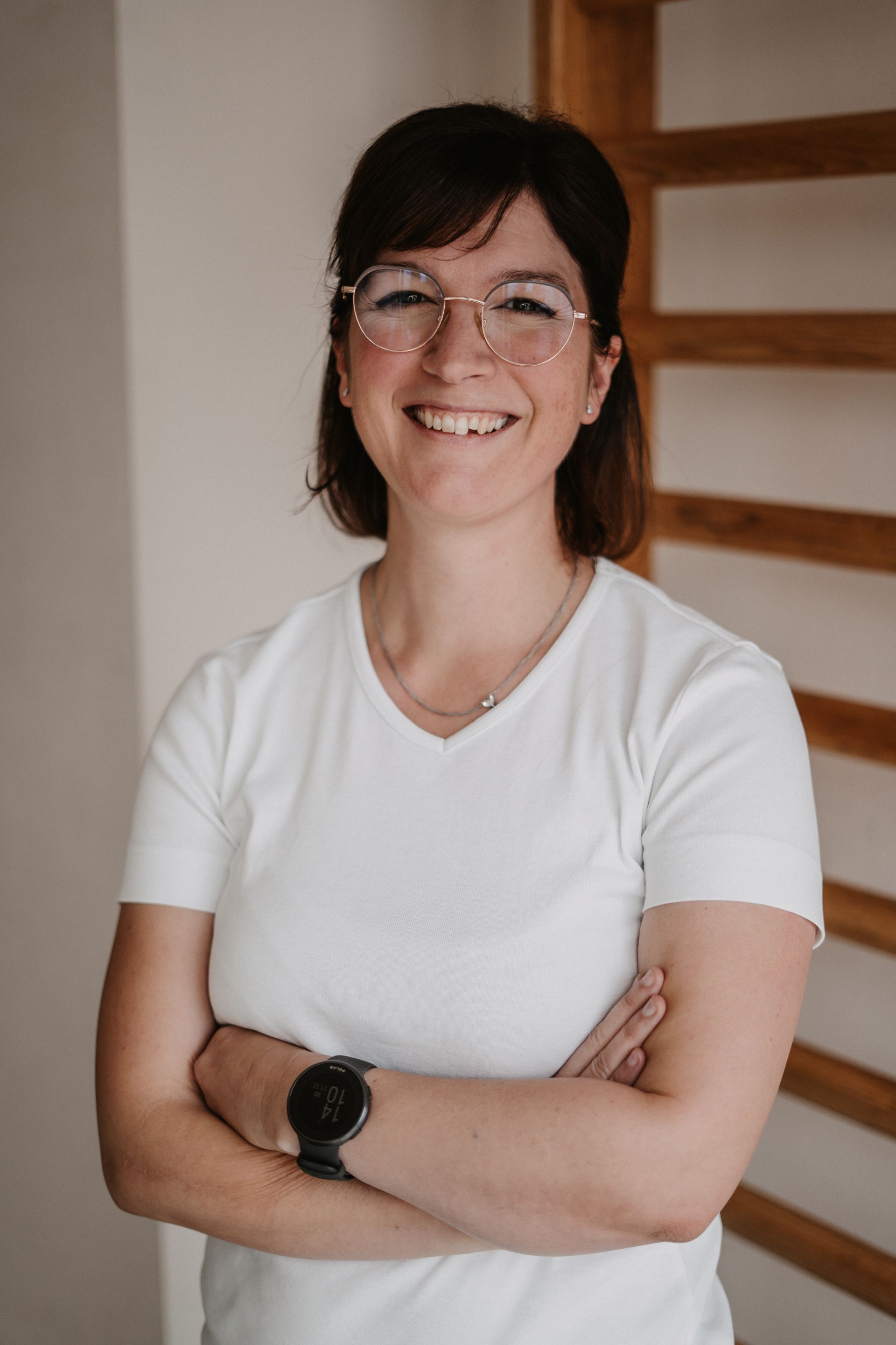 Eine Frau mit Brille und weißem Hemd lächelt und verschränkt die Arme.