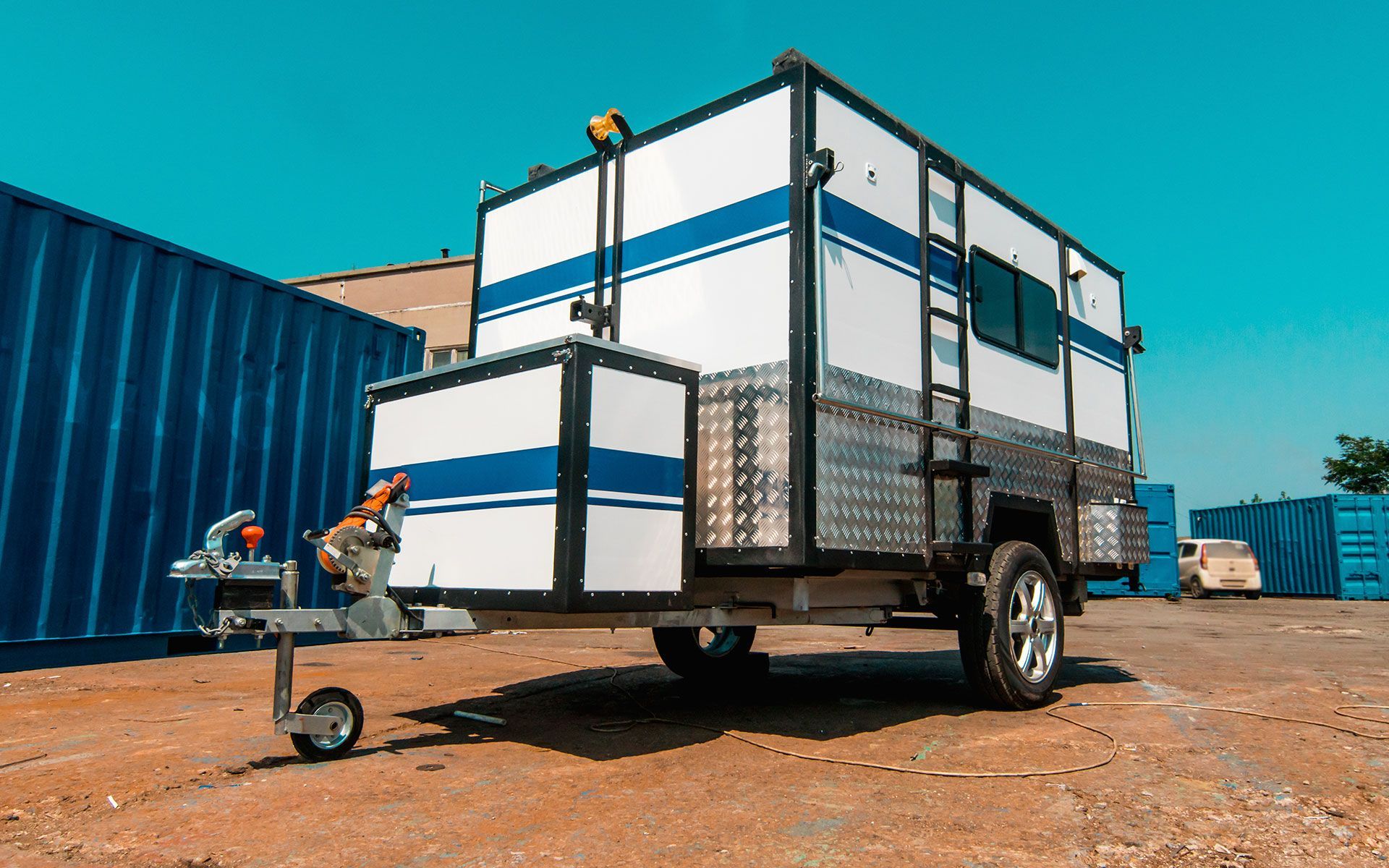 A white and blue trailer is parked in front of a blue container.