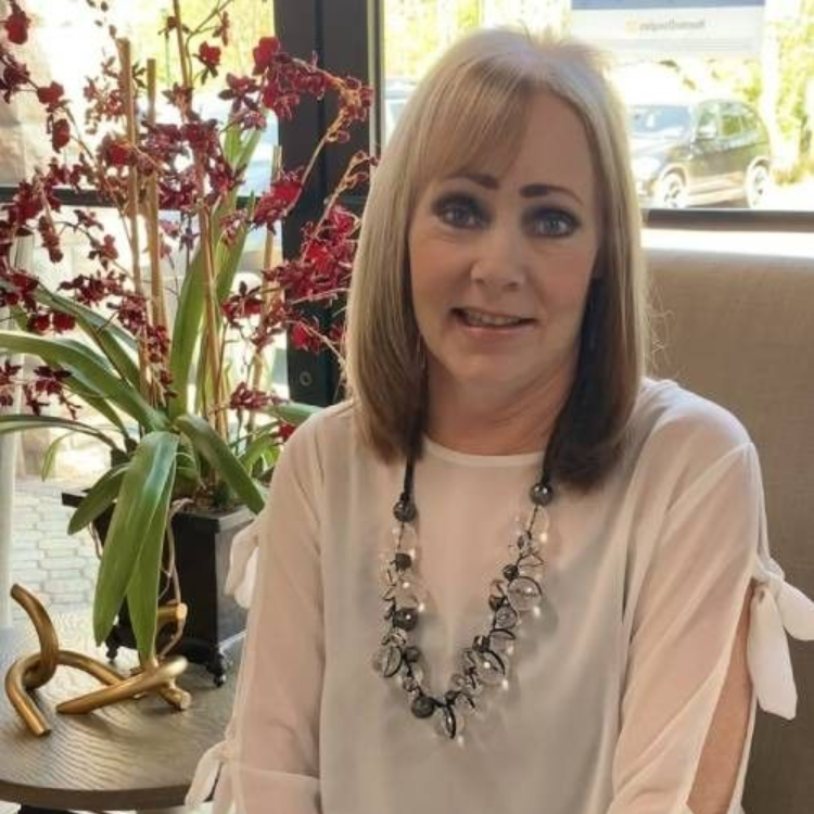 Woman with blonde hair wearing a white top and necklace, smiling. Indoor setting with a plant.