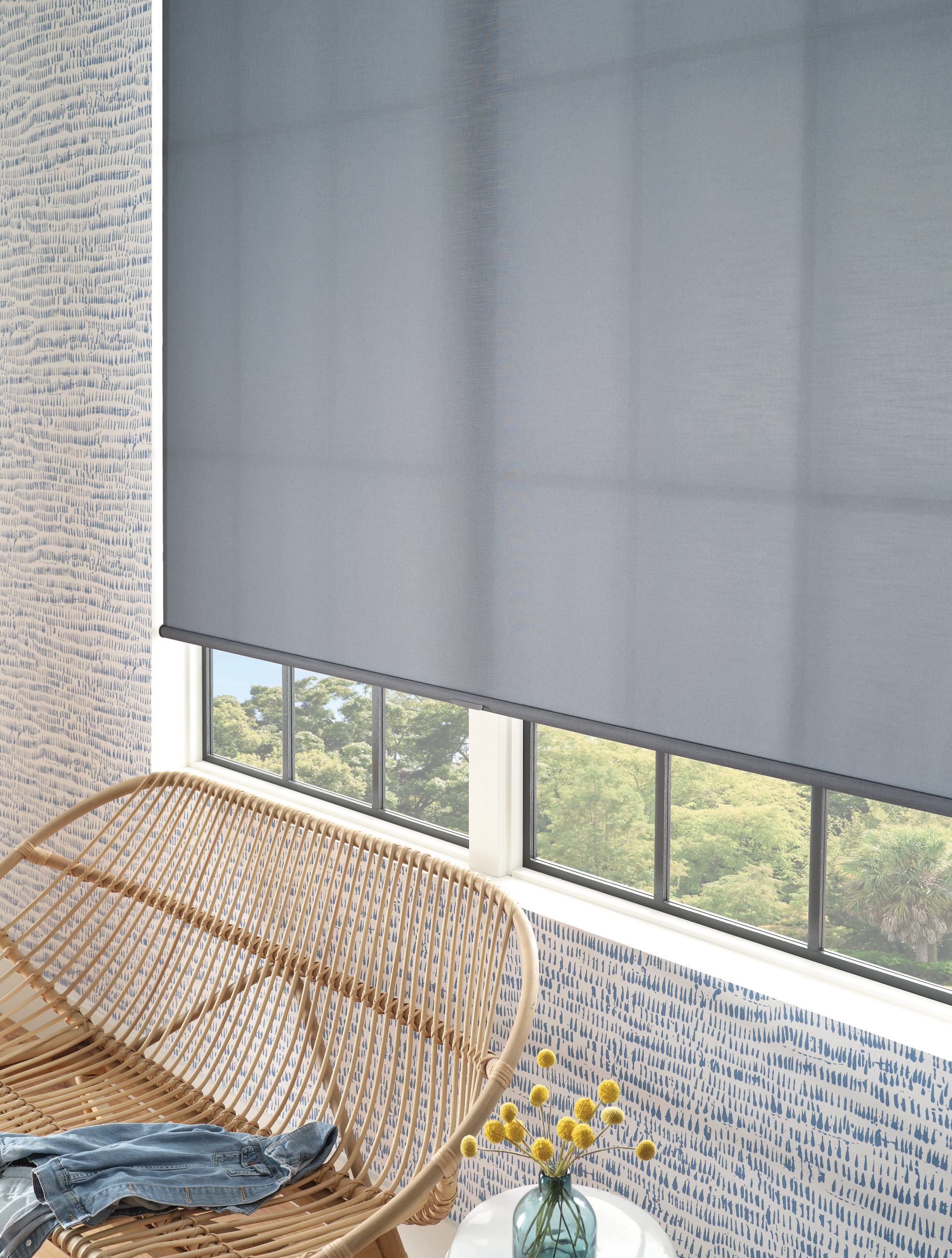 Blue roller shade covering a window with a wicker chair and floral wallpaper.