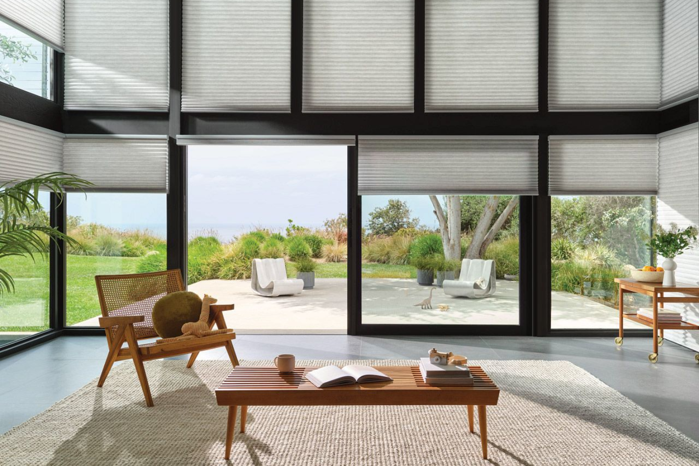 Living room with large windows, open to a patio with seating. Neutral color scheme, blinds.