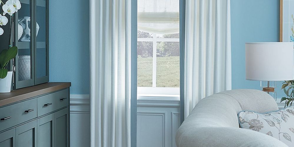 Room with blue walls, white curtains framing a window, and a white sofa with a lamp.