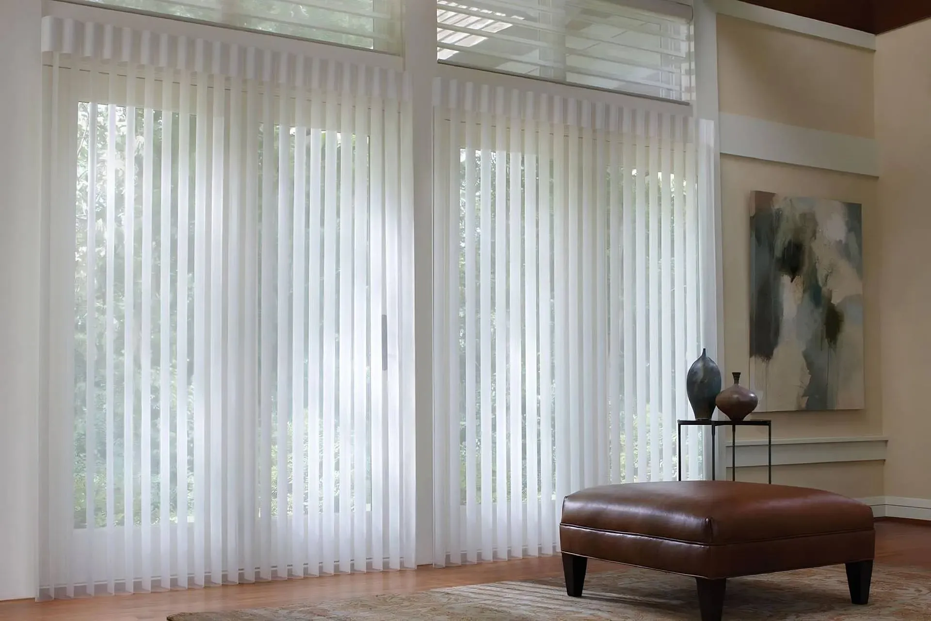 White vertical blinds on large windows; a room with an ottoman, art, and vases.