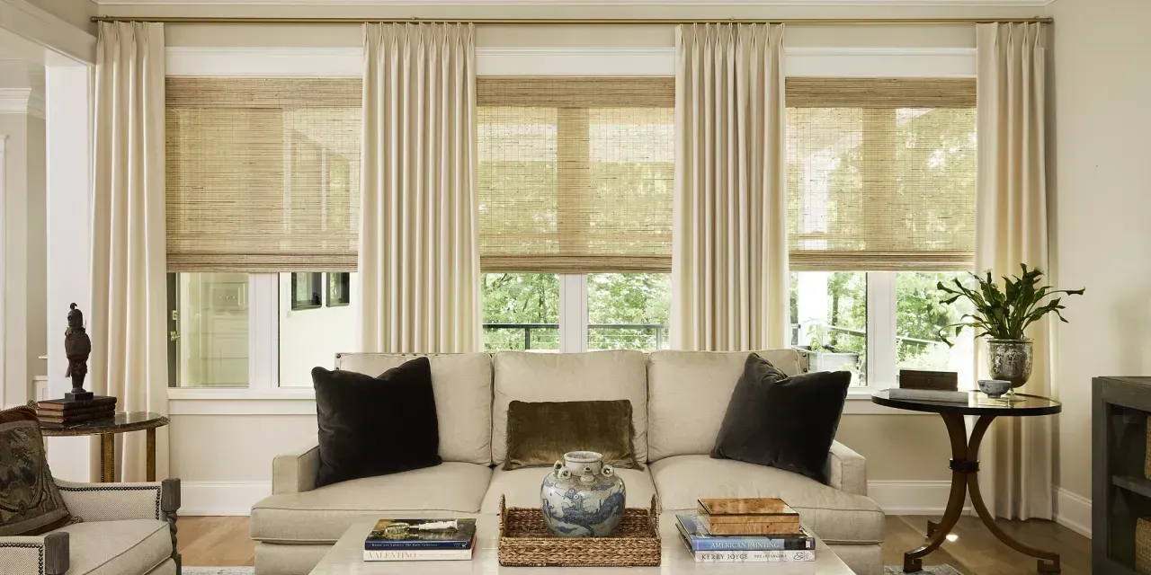 Living room with three windows, each with a woven shade and cream-colored drapes. Beige sofa and small side tables.