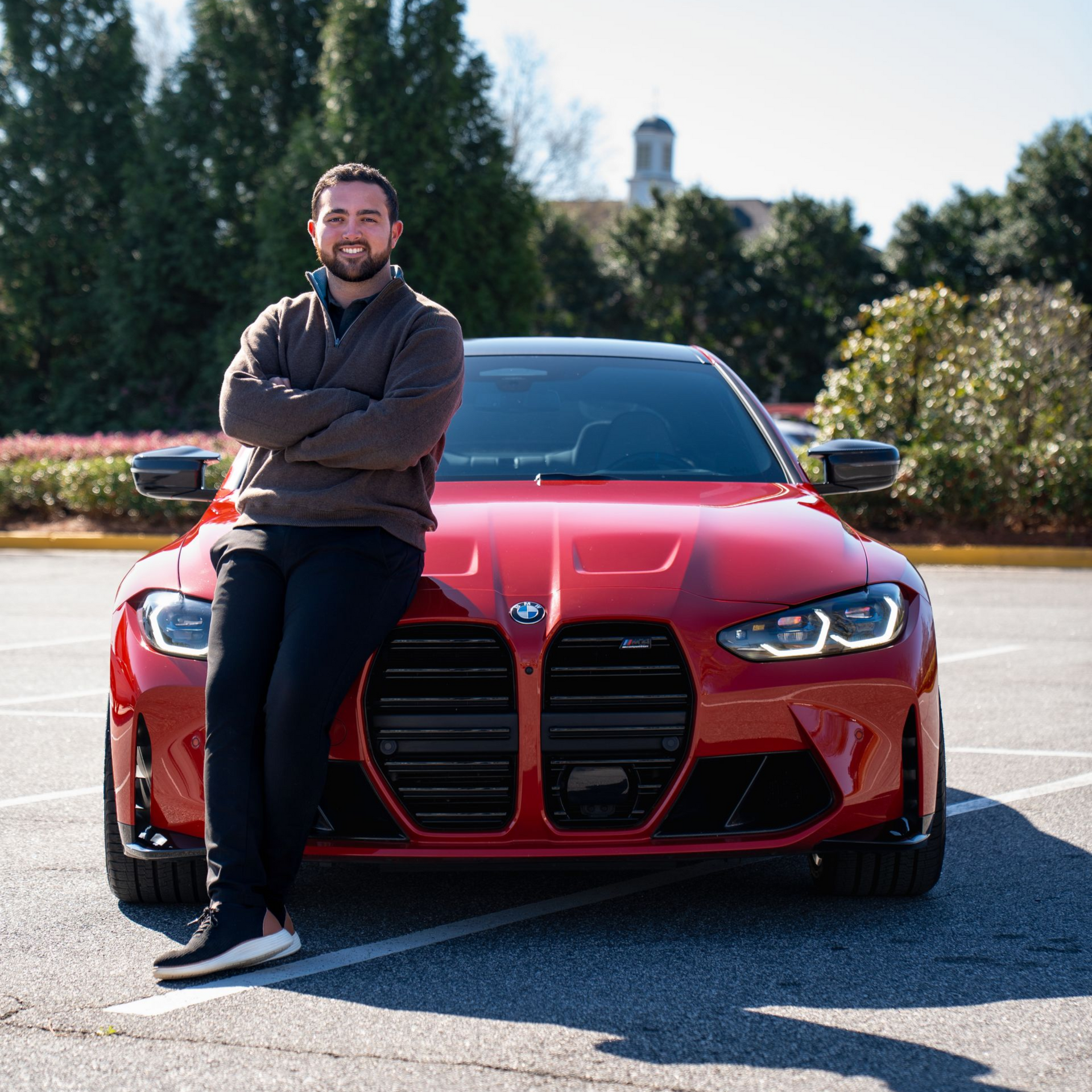 Man leans against a red BMW. He is smiling with arms crossed in a parking lot.
