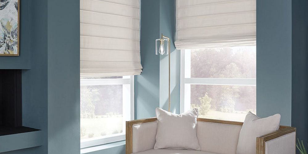 White sofa with pillows in a blue-walled room, two windows with white blinds, and a silver lamp.