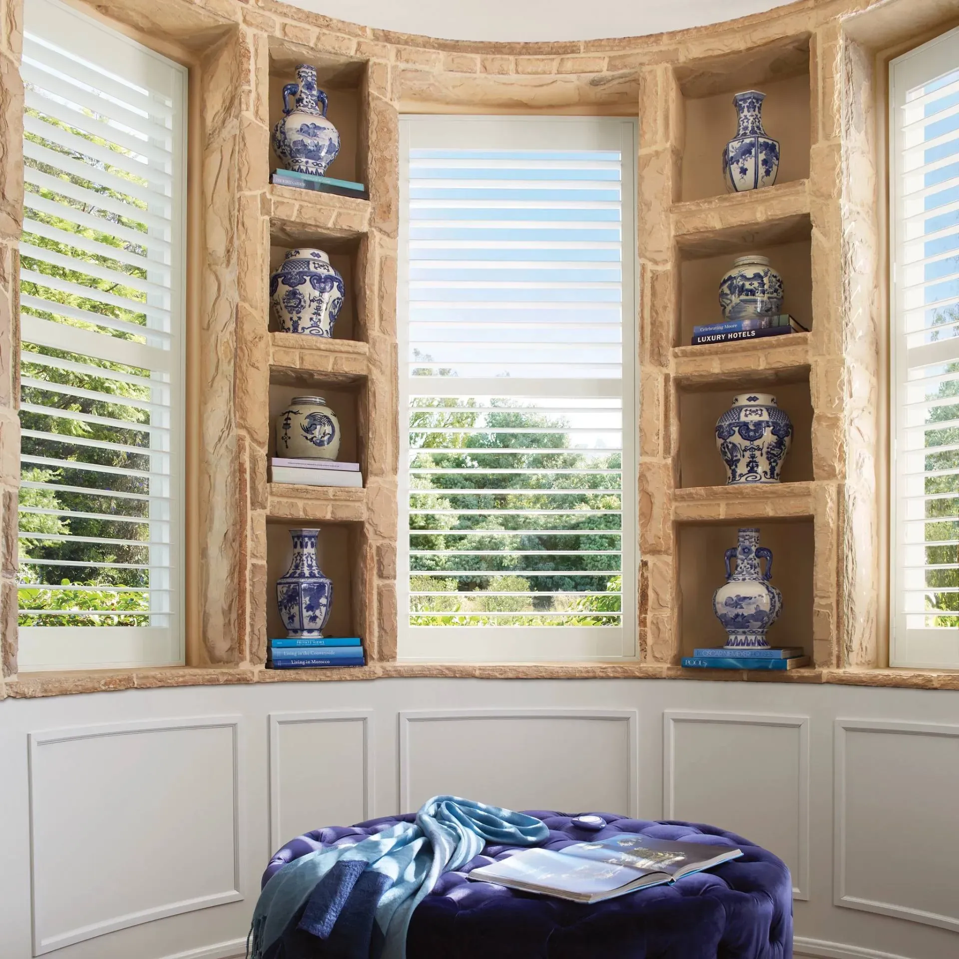Interior with arched stone window, shelves with blue vases, white blinds, and a blue ottoman.
