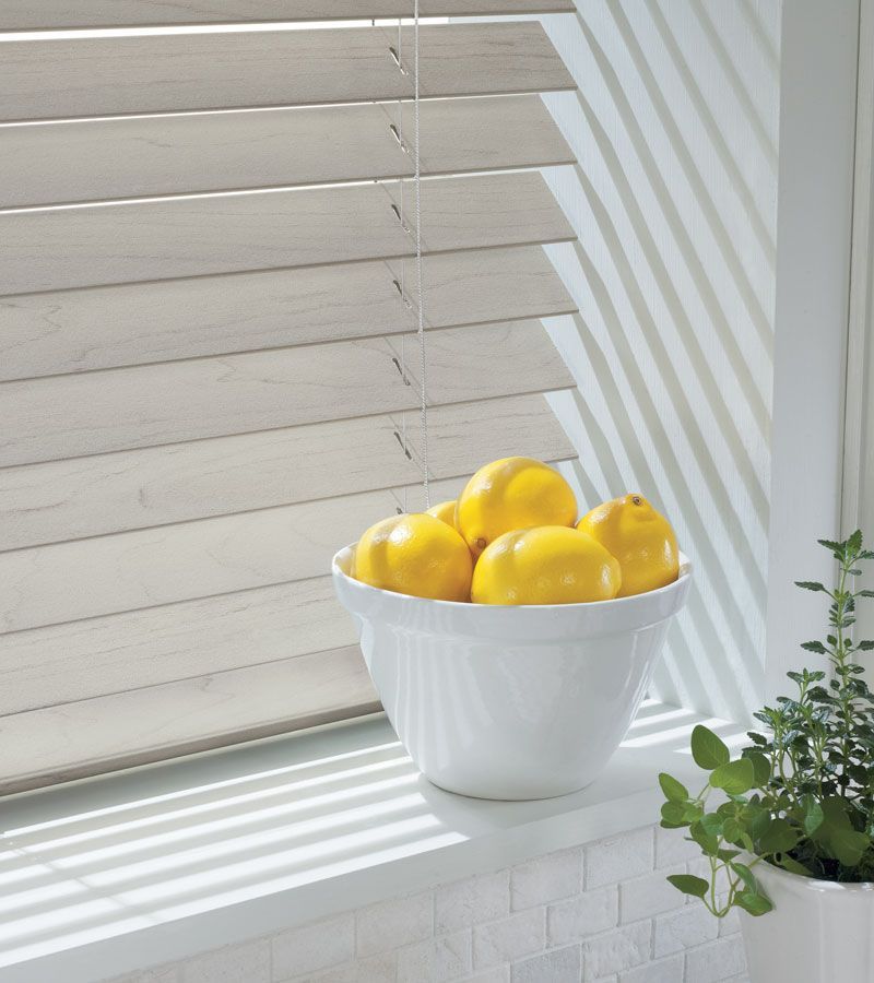 Lemons in a white bowl on a windowsill, beside white blinds and a curtain, bright sunlight.