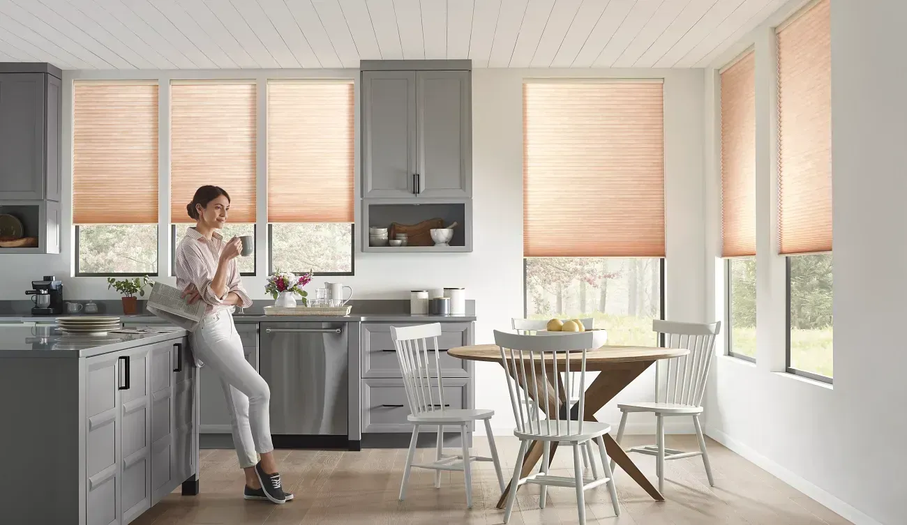 Woman in a kitchen with gray cabinets and peach-colored window shades, leaning against a countertop.