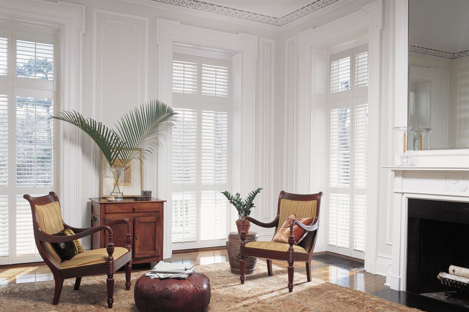 Elegant white room with window shutters, chairs, a fireplace, and a cabinet.