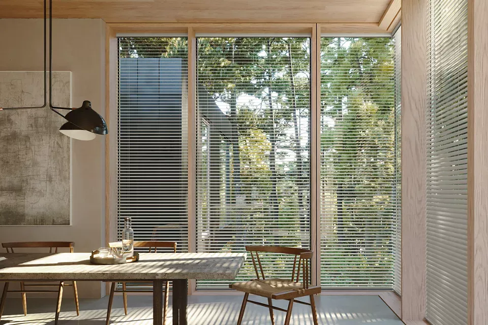 Dining room with large window, wooden table, and chairs. Blinds filter sunlight, with view of trees outside.