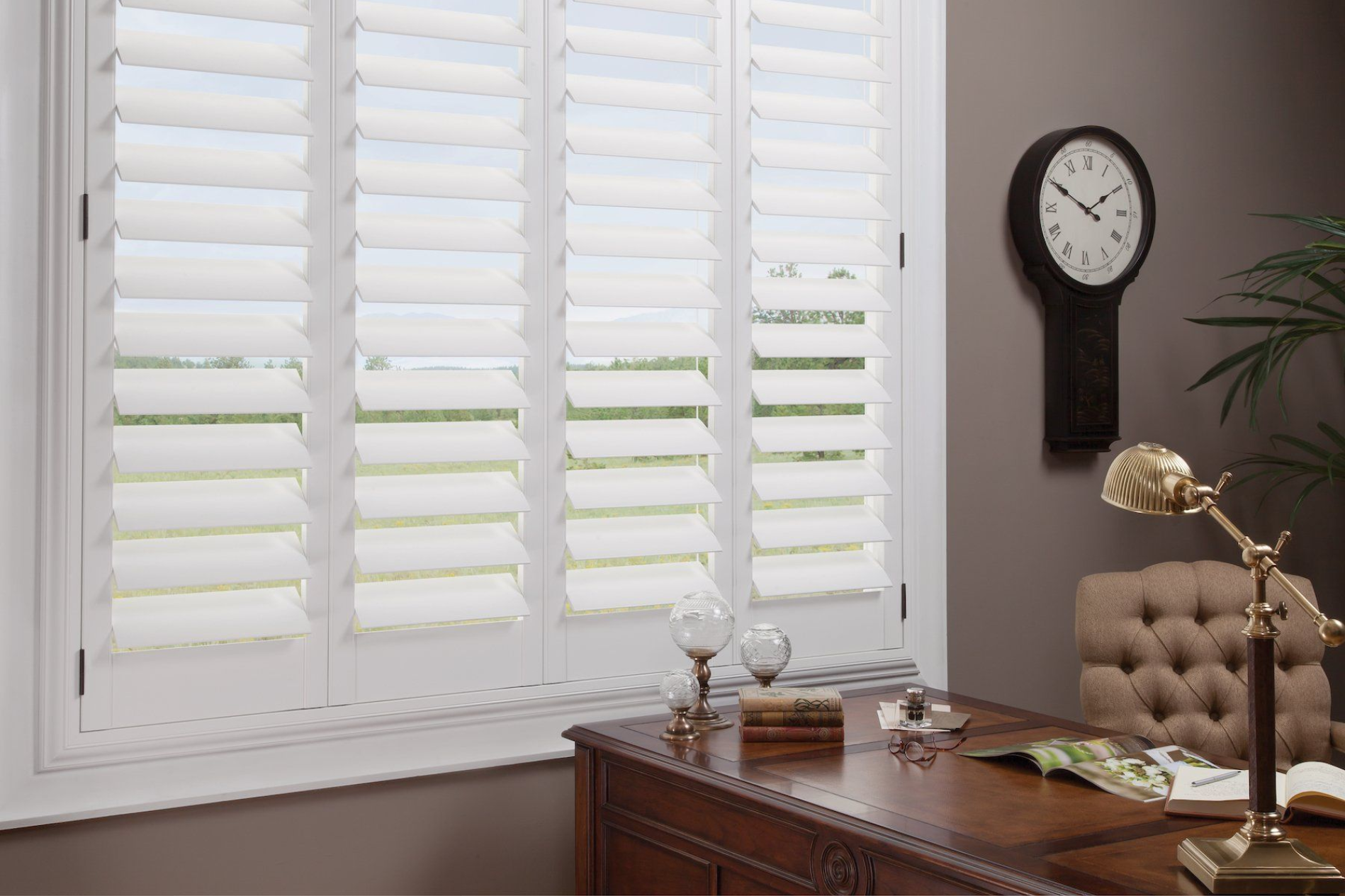 White shuttered window in a room with a desk, clock, and lamp.