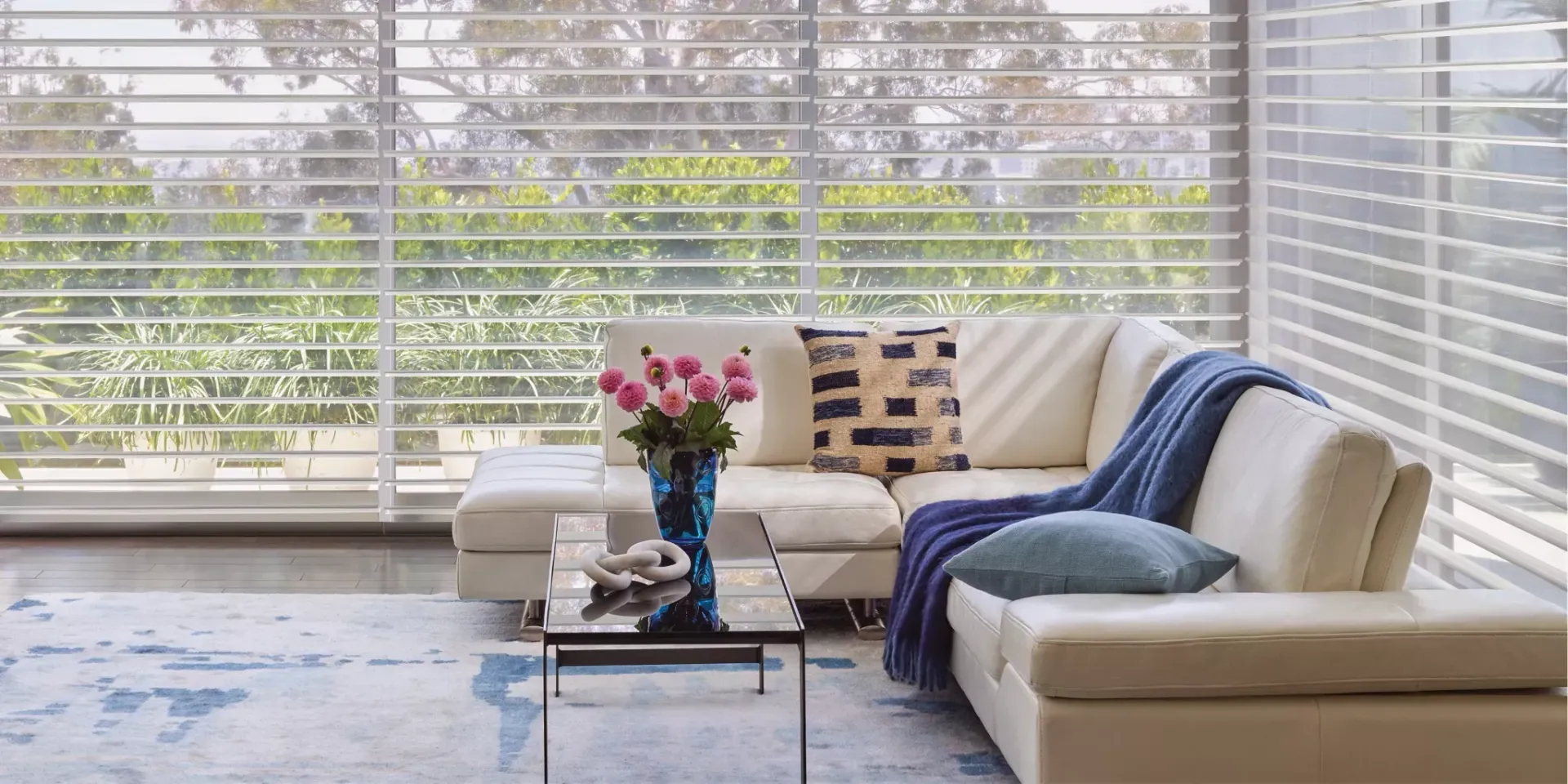 Living room with a white sectional sofa, a blue rug, and large windows with blinds.