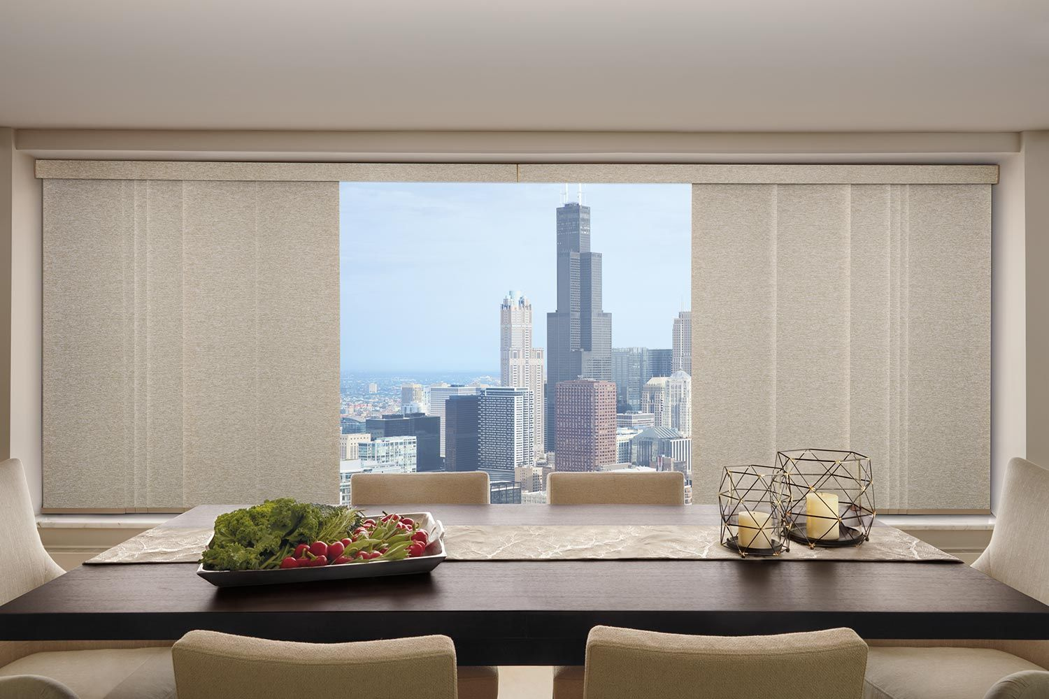 Dining room with a table set for a meal, large window with city view, and neutral-toned panel blinds.