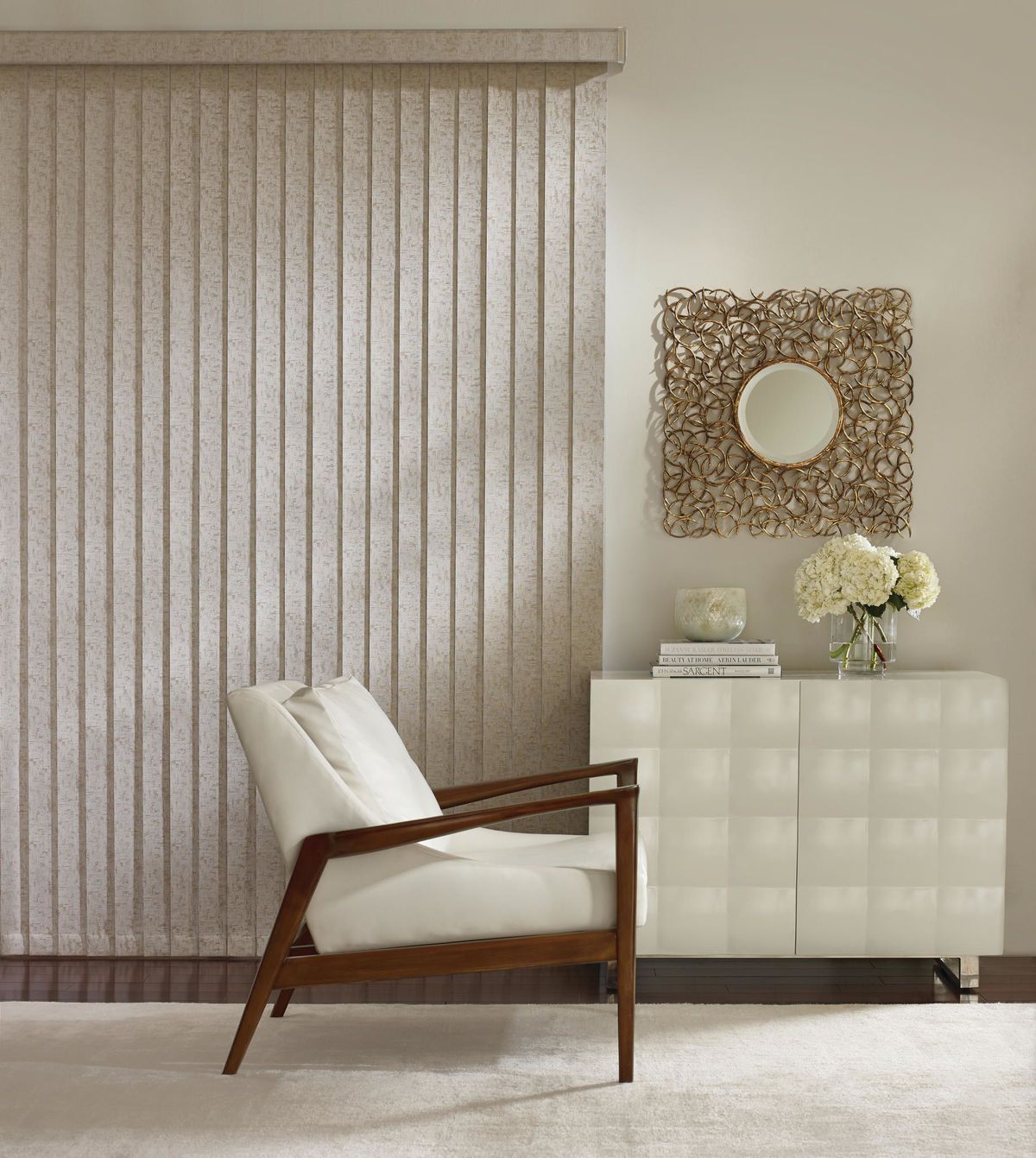 A neutral-toned room with a vertical blind, armchair, white cabinet, mirror, and flowers.