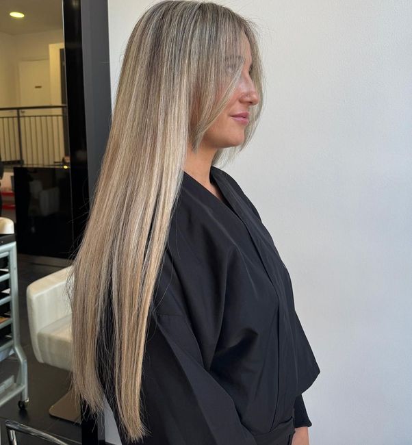 Vista laterale di una donna con lunghi capelli biondi, avvolta in una mantella nera da parrucchiere, all’interno, davanti a una parete bianca.