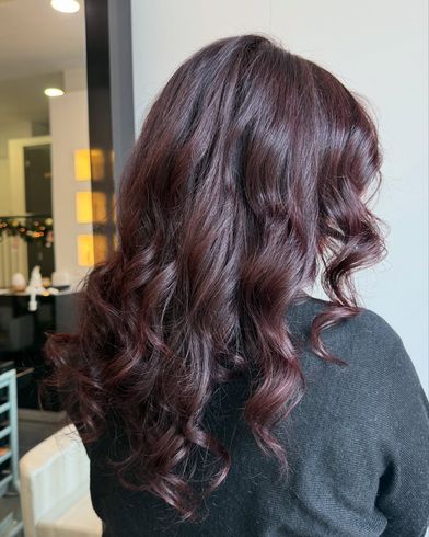 Capelli mossi color bordeaux, acconciati in morbidi ricci, visti da dietro su uno sfondo che ricorda un salone da parrucchiere