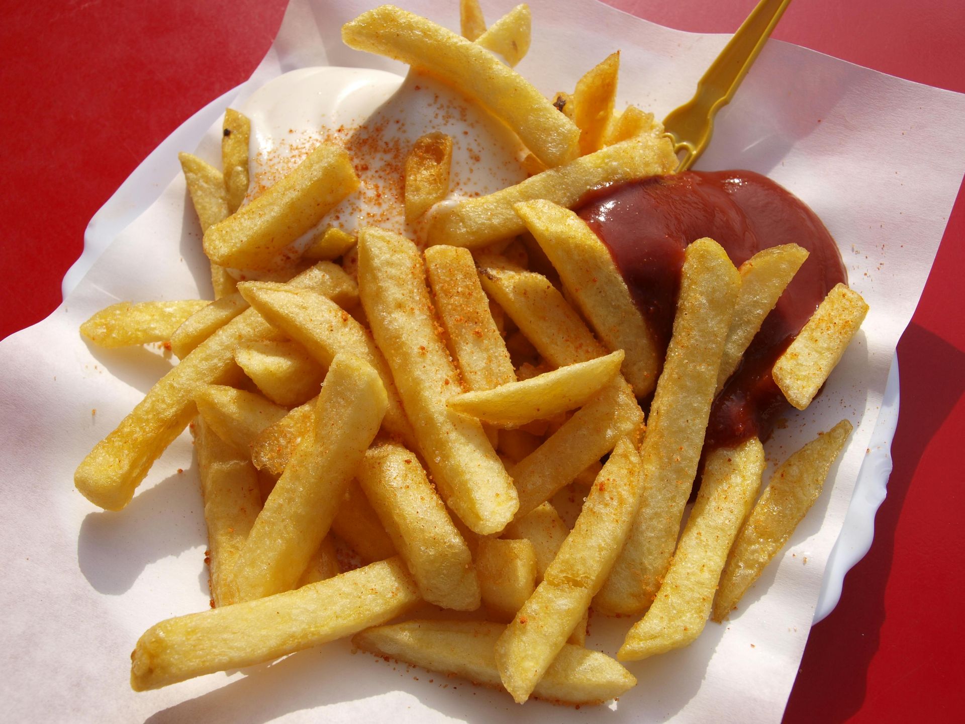 A serving of golden french fries on a paper-lined plate, topped with a dollop of mayonnaise, ketchup, and a plastic fork.