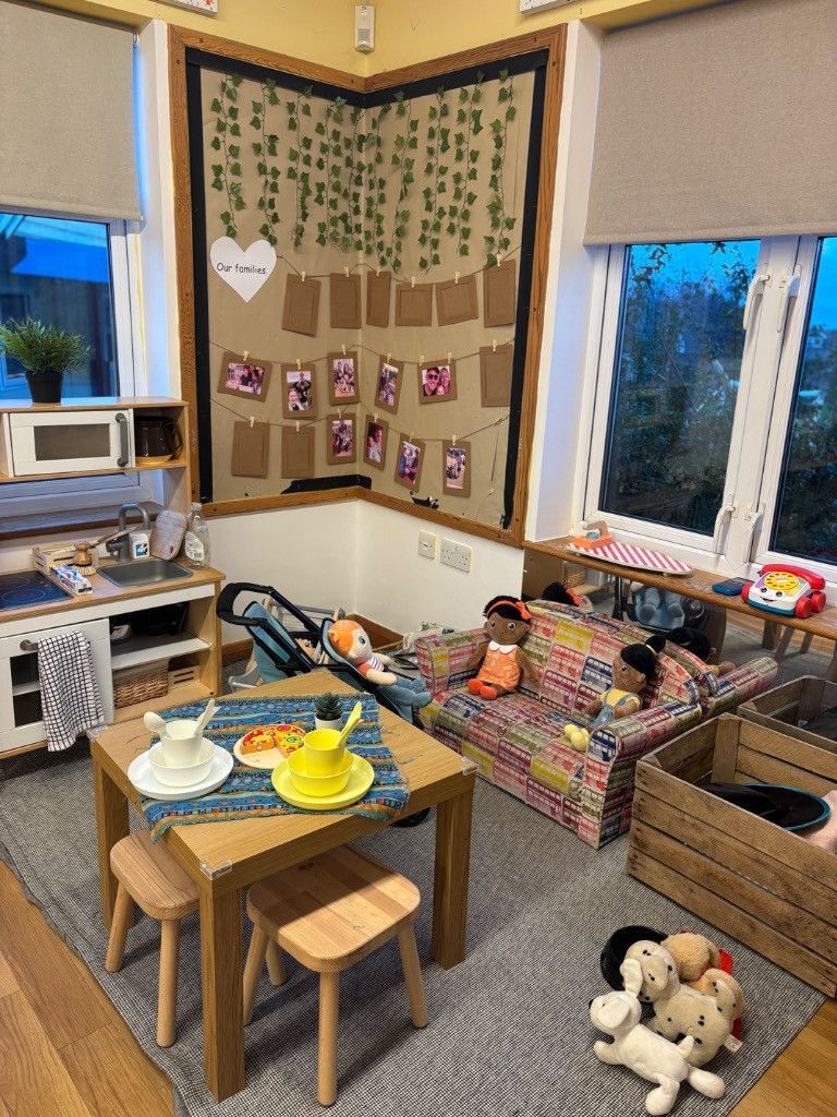 A child's play area with a kitchen, table, sofa, and wall display. Soft toys on the floor.