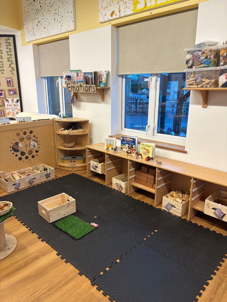 Playroom interior: shelves with toys, window, black floor mat, wooden blocks, and a small patch of green.